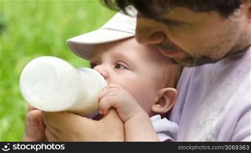 Close-up shot of a young father feeding his baby son from the bottle in the park on a bright summer day