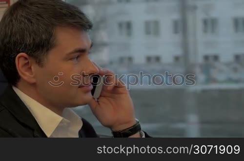 Close-up shot of a young handsome businessman talking on the phone during train ride. City view in the window. Keeping in touch in business trip