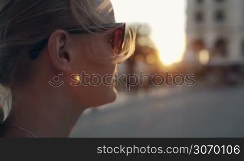 Close-up shot of a young woman in sunglasses looking into distance during sunset, then she turning, smiling and looking away again