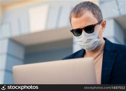 Close up shot of adult man wears sunglasses and medical mask, reads news online on laptop computer, finds out symptoms of coronavirus, has online communication during quarantine and self isolation