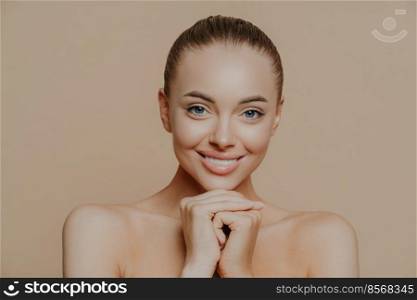 Close up shot of adult woman with fresh daily makeup, smiles toothily and keeps hands under chin, stands half naked indoor, feels refreshed after cosmetic procedures. Beauty and face care concept