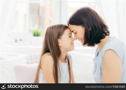 Close up shot of affectionate young mother and her little daughter touches noses and enjoy togetherness, pose against white background and bedroom interior, smile gladfully. Motherhood concept
