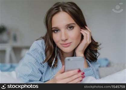 Close up shot of attractive brunette young woman downloads songs from network, uses smart phone for entertainment, dressed in nightclothes, rests in bed, has lazy day, looks seriously at camera