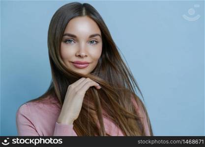 Close up shot of attractive female model shows her sensuality and feminine features, has healthy glowing skin, long dark hair, looks with calm face expression, isolated on blue wall. Beauty and health