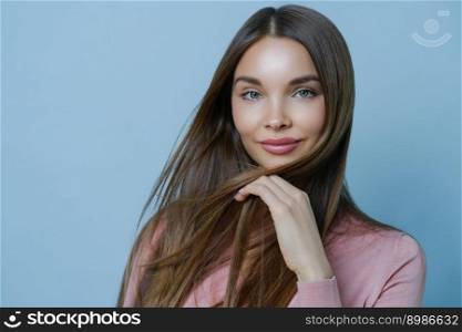 Close up shot of attractive female model shows her sensuality and feminine features, has healthy glowing skin, long dark hair, looks with calm face expression, isolated on blue wall. Beauty and health