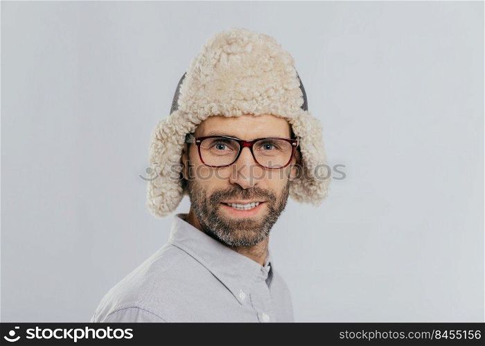 Close up shot of attractive male with stubble, wears glasses and fur hat, prepares for winter, has gentle smile models against white background. Masculine guy has blue eyes thick bristle stands indoor
