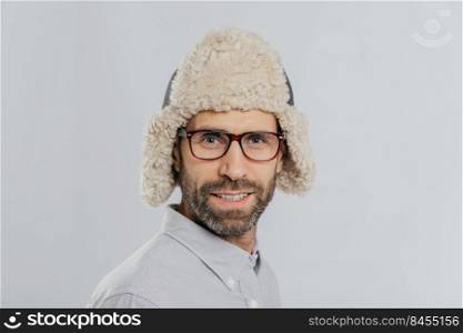 Close up shot of attractive male with stubble, wears glasses and fur hat, prepares for winter, has gentle smile models against white background. Masculine guy has blue eyes thick bristle stands indoor