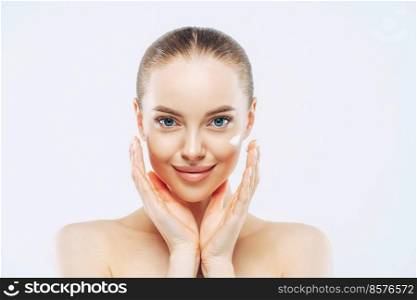 Close up shot of attractive naked young woman with combed hair, applies face cream or lotion, touches face, has natural makeup, poses against white background, looks after skin. High resolution