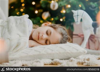 Close up shot of attractive small child lies on floor, sleeps near New Year or Christmas tree, warms with white knitted sweater, has pleasant dreams in evening. Children, winter holidays, rest concept