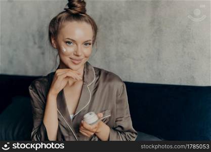 Close up shot of attractive young female model in brown silk pajama sitting on bed in morning after waking up with healthy fresh skin holding cream container in hand and applying cosmetic product. Attractive young female model in brown silk pajama sitting on bed with face cream