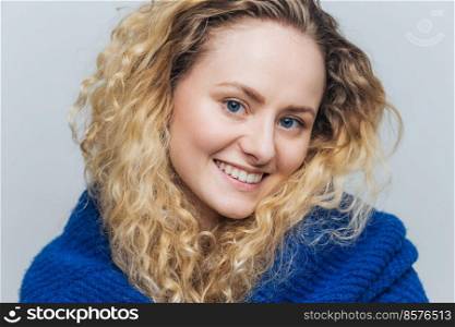 Close up shot of attractive young woman with light crisp hair, blue eyes and shining smile, looks positively at camera, being in good mood after walk outdoor, isolated over light blue background