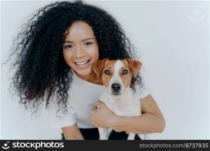 Close up shot of beautiful happy Afro woman with bushy curly hair, embraces favourite dog and have fun together at home, expresses love to jack russell terrier puppy, isolated over white background