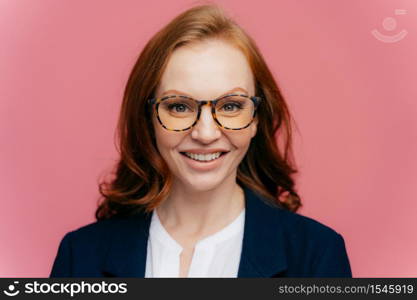 Close up shot of beautiful redhead gorgeous woman in optical glasses, has toothy smile, dressed in elegant clothes, comes on business meeting, happy to sign contract, isolated over rosy background