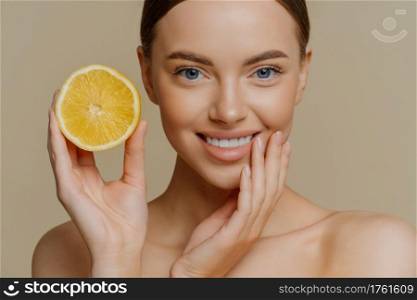Close up shot of beautiful tender woman smiles gently has healthy well groomed skin holds slice of lemon uses citrus fruit for beauty and health stands shirtless isolated over brown background
