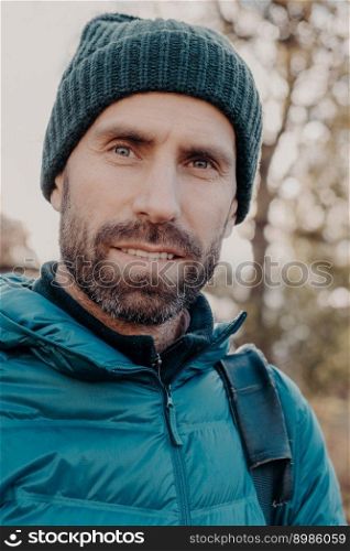 Close up shot of blue eyed man has dark bristle, looks seriously at camera, wears hat and jacket, poses outside, walks in open air, has pleasant appearance. People, lifestyle, masculinity concept