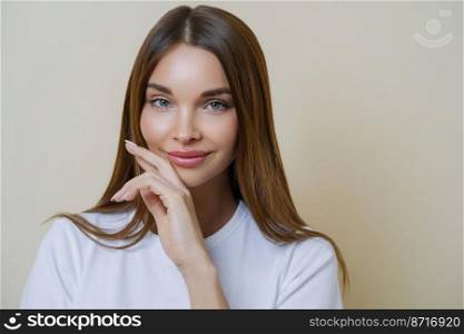 Close up shot of charming dark haired woman keeps hand on cheek, dressed in casual white t shirt, has minimal makeup, poses indoor isolated over beige background. People, beauty, wellness concept
