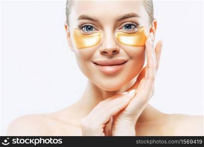 Close up shot of charming tender young woman applies gold collagen patches on fresh facial skin, touches face, has makeup, well cared body, stands against white background. Under eye treatment.