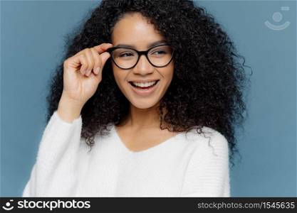 Close up shot of cheerful woman wears glasses, keeps hand on frame, looks gladfully aside, notices something awesome in distance, dressed in casual white jumper, models against blue background