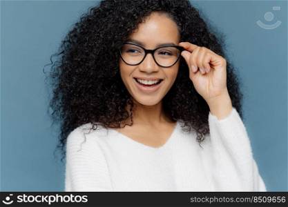 Close up shot of cheerful woman wears glasses, keeps hand on frame, looks gladfully aside, notices something awesome in distance, dressed in casual white jumper, models against blue background