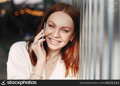 Close up shot of cheerful woman with brown hair, has friendly mobile conversation, has make up, toothy smile, dressed in elegant clothes, has spare time, talks with service operator via app.