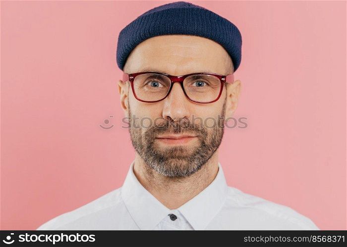 Close up shot of confident male employee with thick beard and mustache, wears transparent glasses, hat, white shirt, stands against pink background, has attentive gaze at camera. Facial expressions