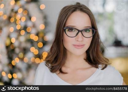 Close up shot of dark haired pretty woman wears eyewear, has serious expression at camera, poses against decorated New Year tree, has appealing look. Beautiful female in spectacles stands indoor