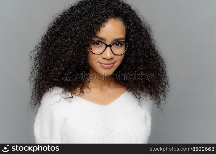 Close up shot of dark skinned female has satisfied expression, bushy curly hair, wears spectacles and white jumper, looks straightly at camera, models over grey background. Natural beauty concept