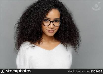 Close up shot of dark skinned female has satisfied expression, bushy curly hair, wears spectacles and white jumper, looks straightly at camera, models over grey background. Natural beauty concept