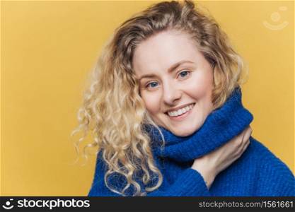 Close up shot of delighted satisfied female model has curly blonde hair, broad smile, demonstrates white perfect teeth, wears blue knitted sweater, isolated over yellow background. Happiness