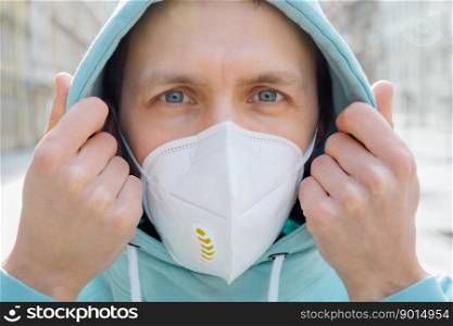Close up shot of European man protects face with respiratory mask, wears hoodie, poses outdoor on street, looks seriosuly at camera, prevents coronavirus or Covid-19, flu. Street during quarantine