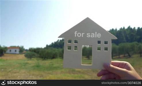 Close-up shot of female hand holding house model with for sale text on the background of woods and blue sky. Real estate business, living in eco place