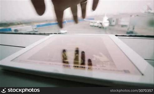 Close-up shot of female hand scrolling photos on pad in airport, takeoff strip with plane in background