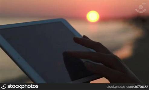 Close-up shot of female hands typing on tablet PC on background of sunset over sea
