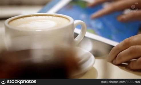 Close-up shot of female hands using touch pad with cup of coffee in foreground. Using tablet in cafe or during coffee break