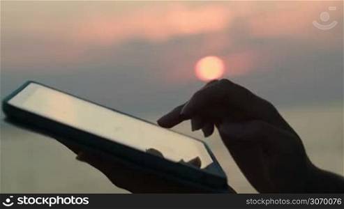 Close-up shot of female hands working on touch pad against background of sea and sky at sunset