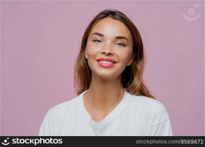 Close up shot of feminine girl smiles gently at camera, wears minimal makeup, has healthy skin, fair hair, feels pleased to get compliment dressed in casual white sweater isolated on purple background