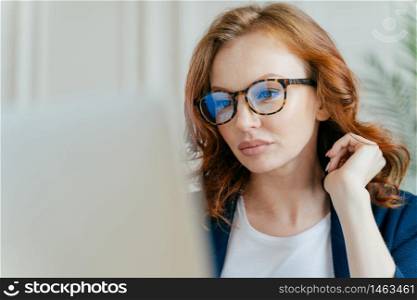 Close up shot of focused female worker completes successful project, makes remote job, focused in monitor of laptop computer, wears optical glasses for vision corrrection, has ginger curly hair