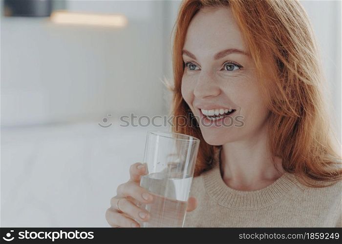 Close up shot of ginger woman drinks pure water from glass enjoys refreshing drink healthy liquid smiles broadly focused into distance wears jumper poses indoor blank space follows healthy lifestyle. Ginger woman drinks pure water from glass enjoys refreshing drink