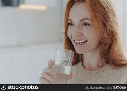 Close up shot of ginger woman drinks pure water from glass enjoys refreshing drink healthy liquid smiles broadly focused into distance wears jumper poses indoor blank space follows healthy lifestyle. Ginger woman drinks pure water from glass enjoys refreshing drink