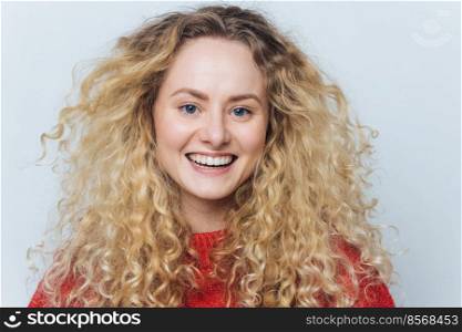 Close up shot of glad adorable woman with curly hair, healthy skin, broad smile and white perfect teeth, happy to spend free time with best friends in funny company, poses against white studio wall