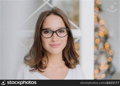 Close up shot of good looking young female model wears spectacles for good vision, looks happily at camera, has pleased expression, charming smile, poses against decorated Christmas tree indoor