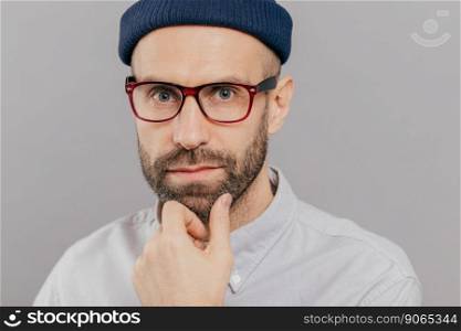 Close up shot of handsome bearded young man holds chin, looks seriously directly at camera, thinks about something, wears spectacles, white clothes, isolated over grey background. Indoor shot