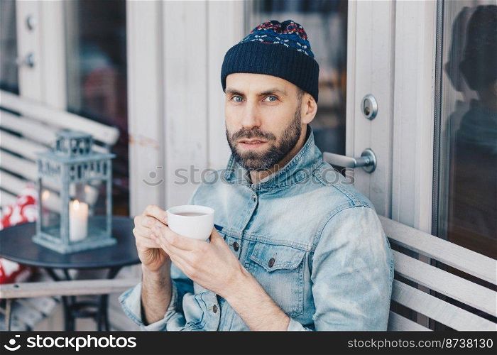 Close up shot of handsome serious bearded male, looks directly into camera with blue eyes and confident expression, drinks hot beverage, enjoys spare time. People, drinks, lifestyle concept.