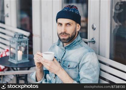 Close up shot of handsome serious bearded male, looks directly into camera with blue eyes and confident expression, drinks hot beverage, enjoys spare time. People, drinks, lifestyle concept.