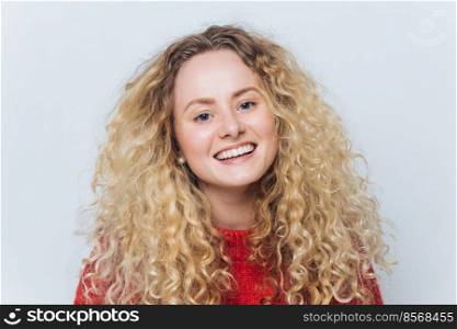 Close up shot of happy attractive woman with broad shining smi≤, bushy blonde curly hair, poses against white background, satisfied to pass examination session successfully. Peop≤, beauty.