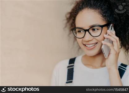 Close up shot of happy dark skinned woman with Afro hairstyle talks via mobile phone, smiles gently, wears spectacles and white t shirt, gets good news, isolated on brown background, empty space