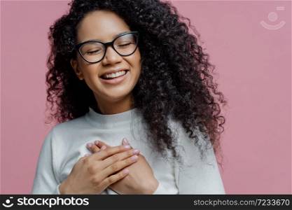Close up shot of happy sincere Afro American woman keeps hands pressed to chest, expresses sincere feelings, makes gratitude gesture, feels thankful for help, smiles broadly, wears spectacles