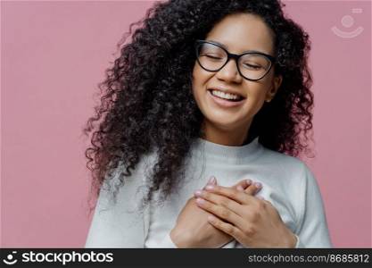 Close up shot of happy sincere Afro American woman keeps hands pressed to chest, expresses sincere feelings, makes gratitude gesture, feels thankful for help, smiles broadly, wears spectacles