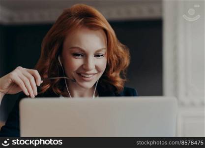 Close up shot of happy young woman freelancer concentrated at laptop computer, watches interesting tutorial webinar, uses earphones. Female entrepreneur has online seminar during remote job.