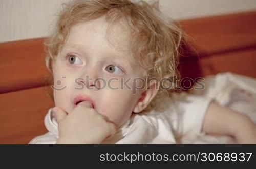 Close-up shot of little boy on bed looking somewhere with his fingers in the mouth